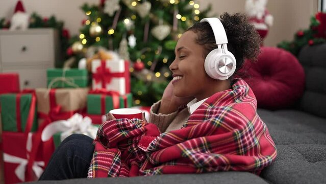 African American Woman Listening To Music Drinking Coffee Sitting By Christmas Tree At Home