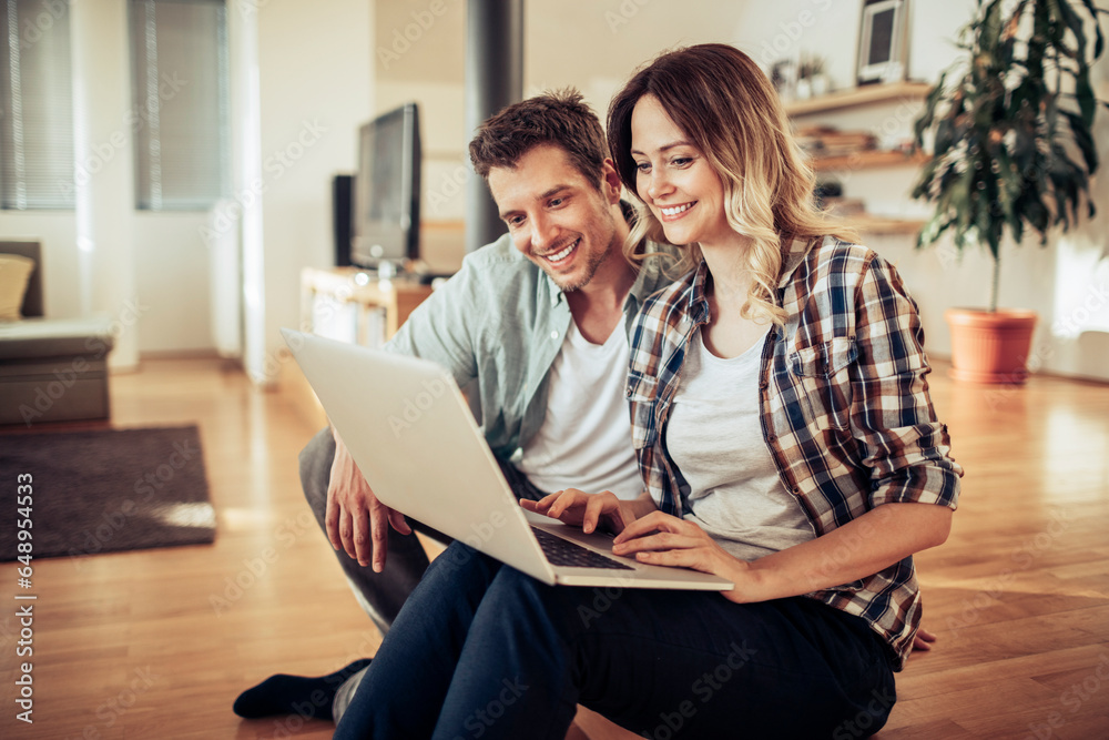 Wall mural Young Caucasian couple using a laptop in the living room at home