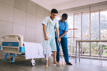 Male patient walking slowly with crutch while nurse supporting at hospital ward.