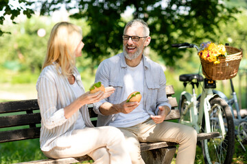 Happy Senior Couple Eating Sandwiches While Sitting On Bench In Park
