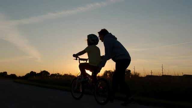Mom teaches daughter to ride a bike. Happy family childhood dream concept of parent and little kids learn to ride a bike silhouette in the park.