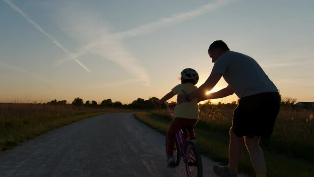 Dad teaches daughter to ride a bike. Happy family childhood dream concept of parent and little kids learn to ride a bike silhouette in the park.  Wide shot