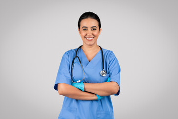 Positive brazilian female nurse posing with crossed arms, smiling at camera, wearing blue coat and...