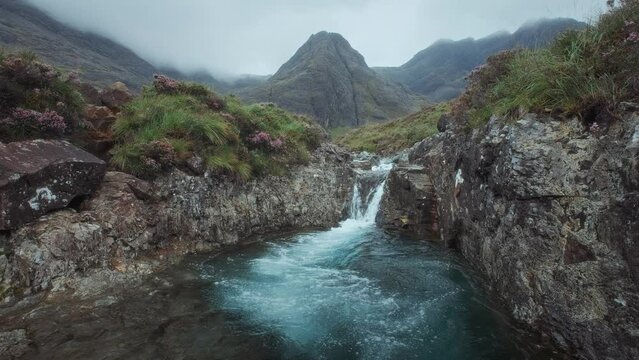 Footage of a morning view of a river with a waterfall and a mountain range covered in clouds. Fairy Pools, Isle of Skye, Scottish Highlands, UK