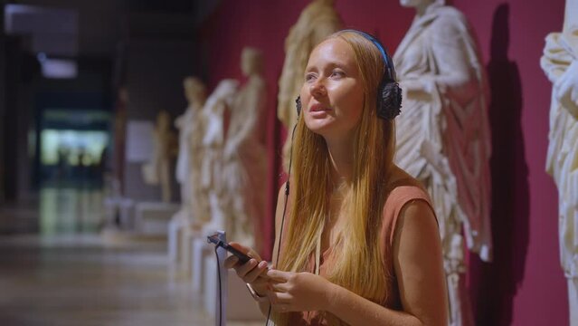 A young woman is portrayed exploring a history museum, attentively listening to an audio guide. The camera captures her inquisitive expressions and genuine interest as she immerses herself in the
