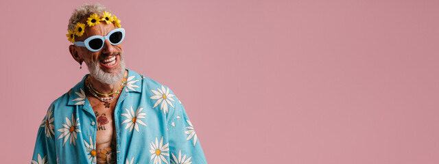 Cool senior man in stylish shirt and floral wreath on head smiling against pink background