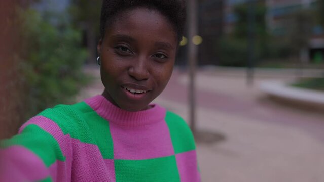 Happy young african woman having a video call with friend using mobile phone app while sitting outdoors over urban background. Screen device view. She is waving hand.