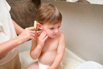 A mother is gently brushing her child hair with a soft brush near the sink in the bathroom. Kid...