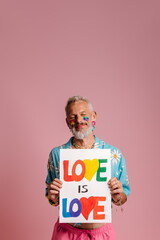Happy mature gay man with rainbow flag make-up showing colorful banner against pink background