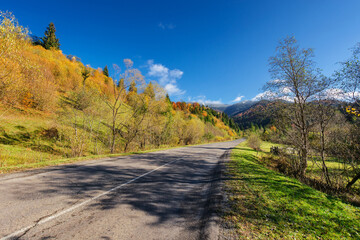 Fototapeta na wymiar old asphalt road through countryside landscape in autumn. scenery with trees on the hill in colorful foliage. clouds on the blue sky above the distant forested mountain