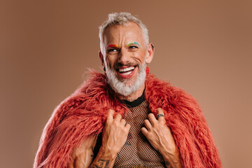Happy mature gay man with rainbow colored eyebrows wearing fluffy coat against brown background