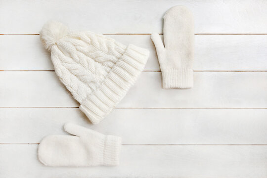 A white knitted hat with a pompom and woolen mittens lie on a white wooden background. Details of winter clothing