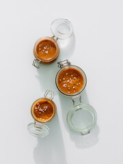 Salted caramel in glass jars, top view. Brown caramel or condensed milk with sea salt crystalls, shoot in bright or hard light. Light neutral background. Vertical