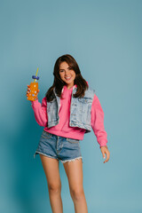 Attractive young woman in denim wear holding bottle with lemonade and smiling against blue background