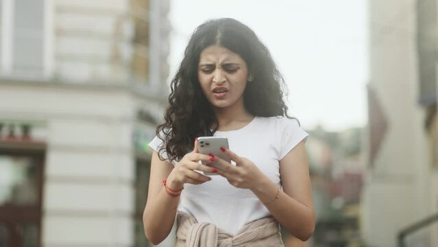 Unhappy young curly woman feeling annoyed when getting message on smartphone at city street Nervous female having problem conflict online outdoors