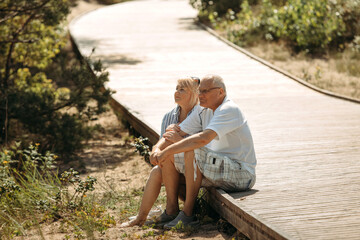 A happy elderly couple is sitting on a wooden path in a pine forest in the dunes and talking