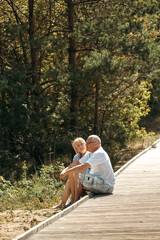 A happy elderly couple is sitting on a wooden path in a pine forest in the dunes and talking