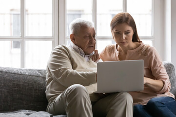 Grownup daughter sit on couch at home with elderly father teach using laptop gadget, smart modern old 80s dad browsing computer with adult millennial girl child, relaxing with device together