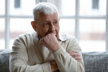 Thoughtful elderly 80s grandfather sit on couch in living room look in distance thinking or pondering, pensive upset mature 70s man feel lonely sad, mourn or yearn, lack communication at home