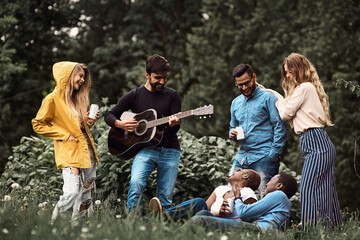 Young man plays acoustic guitar with a mixed-gender and mixed-race group of friends, enjoying leisure time outdoors amidst nature in spring. Outdoor company party. Guitar classes advertisement.