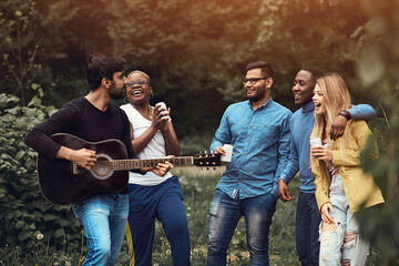 A young man plays the guitar in a multiethnic company of five people. Friends having fun in a park during sunset. Impromptu jam session. Hanging out, get-together. Corporate team building event.