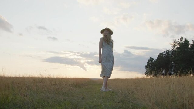 A young girl in a hat spins in a field against the backdrop of a beautiful summer sunset and looks at the camera. Slow motion. Medium shot