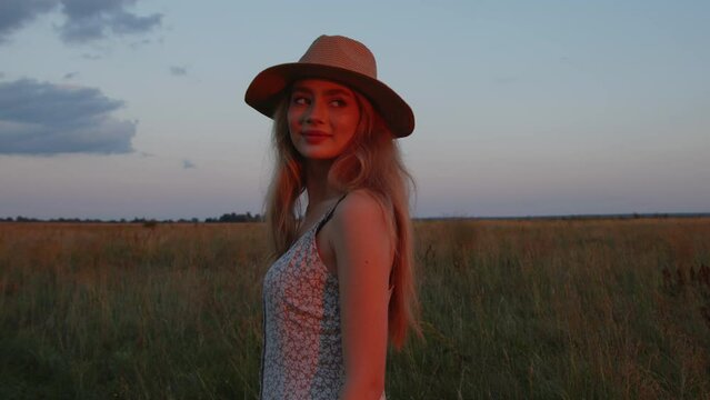 A young girl in a hat runs across a field against the backdrop of a beautiful summer sunset and looks at the camera. Slow motion. Close-up
