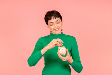 happy woman wearing green casual sweater saving money for rainy day in the piggy bank, investment, saving money, currency, deposit. Indoor studio shot isolated on pink background.