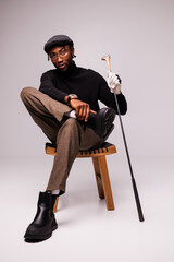 Fashionable african american man in flat cap and eyeglasses holding golf club on chair on grey 
