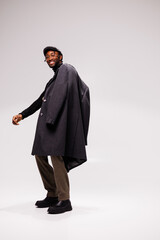 Obraz na płótnie Canvas Cheerful and fashionable african american man in flat cap ad coat posing on grey background
