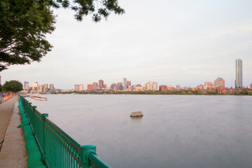 Fototapeta premium The Charles River is running by Boston in the distance and Cambridge on the left. A green metal fence is on the left. A green tree is on the upper left. The river is very tranquil and there is rock.
