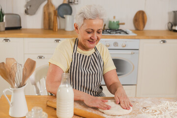 Happy senior woman cooking in kitchen. Stylish older mature gray haired lady grandmother knead...
