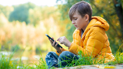 a boy in a yellow jacket sits on the grass in the park, plays a game on his mobile. A 10-year-old child chats outdoors, looks at social networks outside.