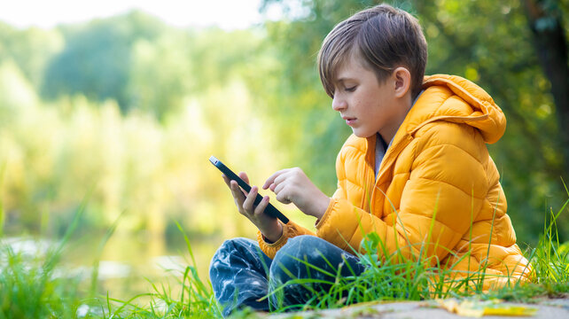 a boy in a yellow jacket sits on the grass in the park, plays a game on his mobile. A 10-year-old child chats outdoors, looks at social networks outside