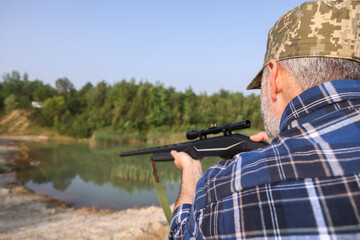 Man with hunting rifle near lake outdoors, back view. Space for text