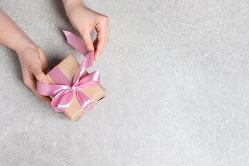 Woman holding gift box with pink bow at light grey table, top view. Space for text