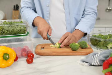 Man cutting fresh broccoli with knife near containers at white marble table in kitchen, closeup. Food storage