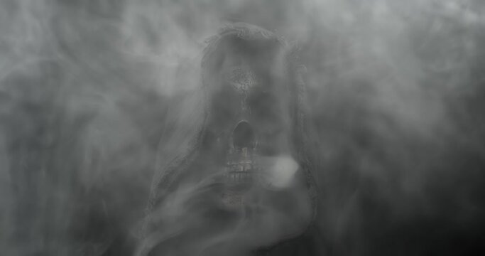 Smoke surrounding mysterious hooded figure with spooky human skull slowly fading into darkness. Scary grim reaper representing death, horror, or frightening Halloween silhouette.