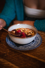 Shot of a young woman scooping a spoonful of an acai bowl with raspberry and granola