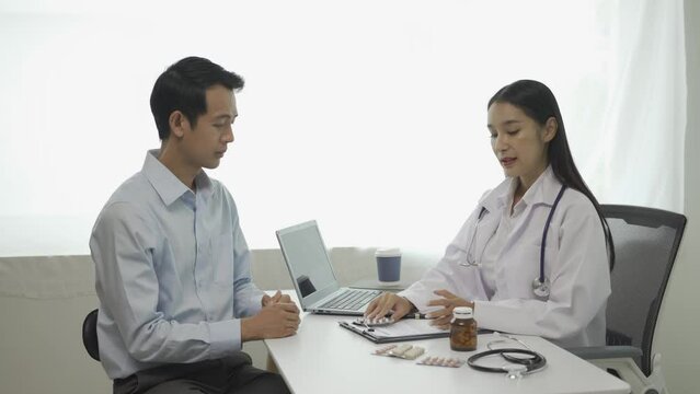 Young Asian doctor in white medical uniform uses clipboard to discuss results or symptoms with male patient sitting at table in clinic to discuss health issues.
