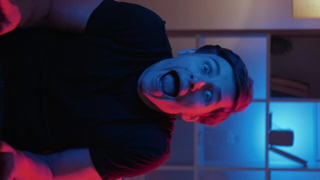 Vertical video. Grimacing man. Meme expression. Silly behavior. Foolish guy making nuts scare face sitting sofa in dark neon light interior looped.