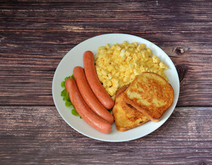A plate of delicious breakfast, boiled sausages, egg scrambled eggs and fried croutons on a dark wooden table.