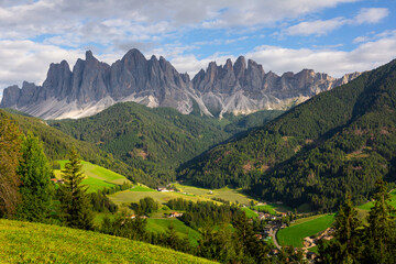 Fototapeta na wymiar Breathtaking view of lush green Val di Funes valley with rustic cottages nestled amidst pristine alpine landscape against backdrop of rugged peaks 