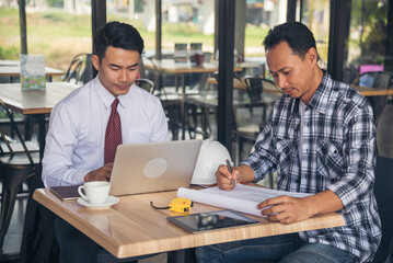 Obraz premium Asian engineer man brainstorming teams partner meeting office desk on construction site. Engineering men building contractor civil worker team meeting together. Men look at construction Blueprint