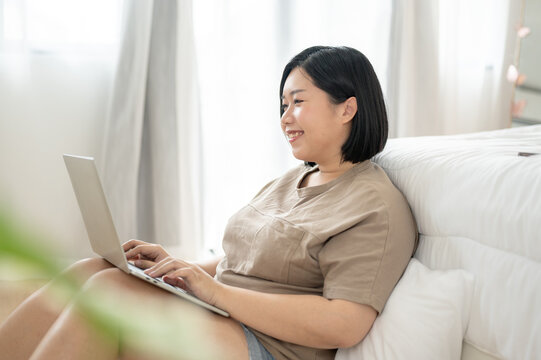 A happy Asian plus-size woman is using her laptop while relaxing in her bedroom. work from home