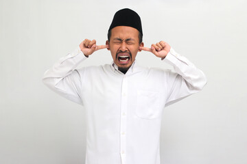 Angry Young Indonesian man, wearing a songkok, peci, or kopiah, covers his ears, feeling the...