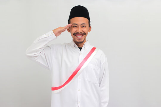 Excited young Indonesian man, wearing red and white sash and either peci or kopiah, is making a salute gesture while celebrating Indonesia's Independence Day on 17 August, isolated on white background