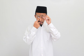 A young Indonesian man, wearing a songkok, peci, or kopiah, is experiencing sore eyes and rubbing...