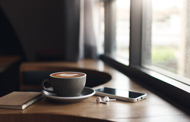 Wireless earphones and mobile with latte coffee and note book on wooden table by bright window, people relax by using mobile phone for entertainment and social media online at cafe