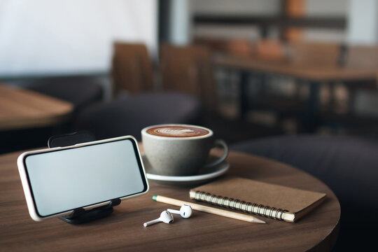 Smart phone with blank screen, wireless earphones and note book on wooden table, freelancer working part time work and meeting online vie smart phone in cafe
